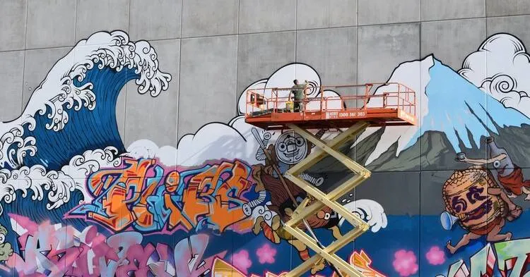 Large mural of a Japanese-style great wave, Mount Fuji and graffiti lettering on the big wall in Braddon, Canberra, painted from an orange scissor lift