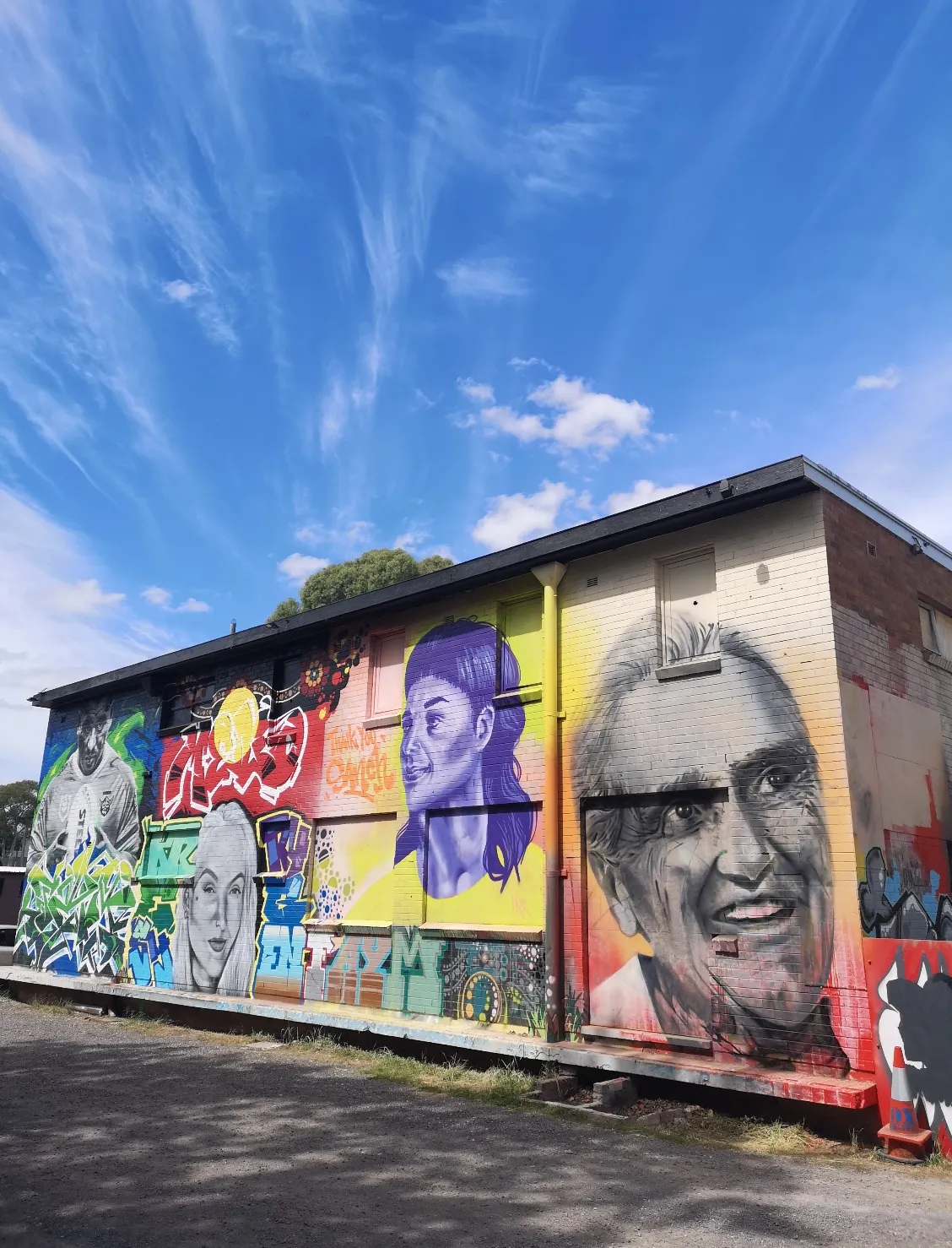 Building side wall carrying four large portrait murals under a blue sky, including an elderly woman and a young man