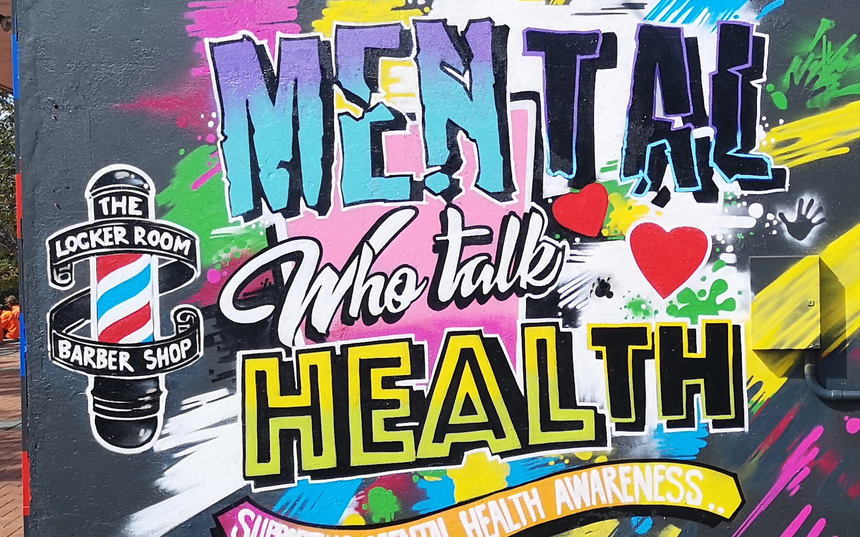 Wide daytime shot of a lettering mural reading 'Mental Health, who talk' outside The Locker Room Barber Shop