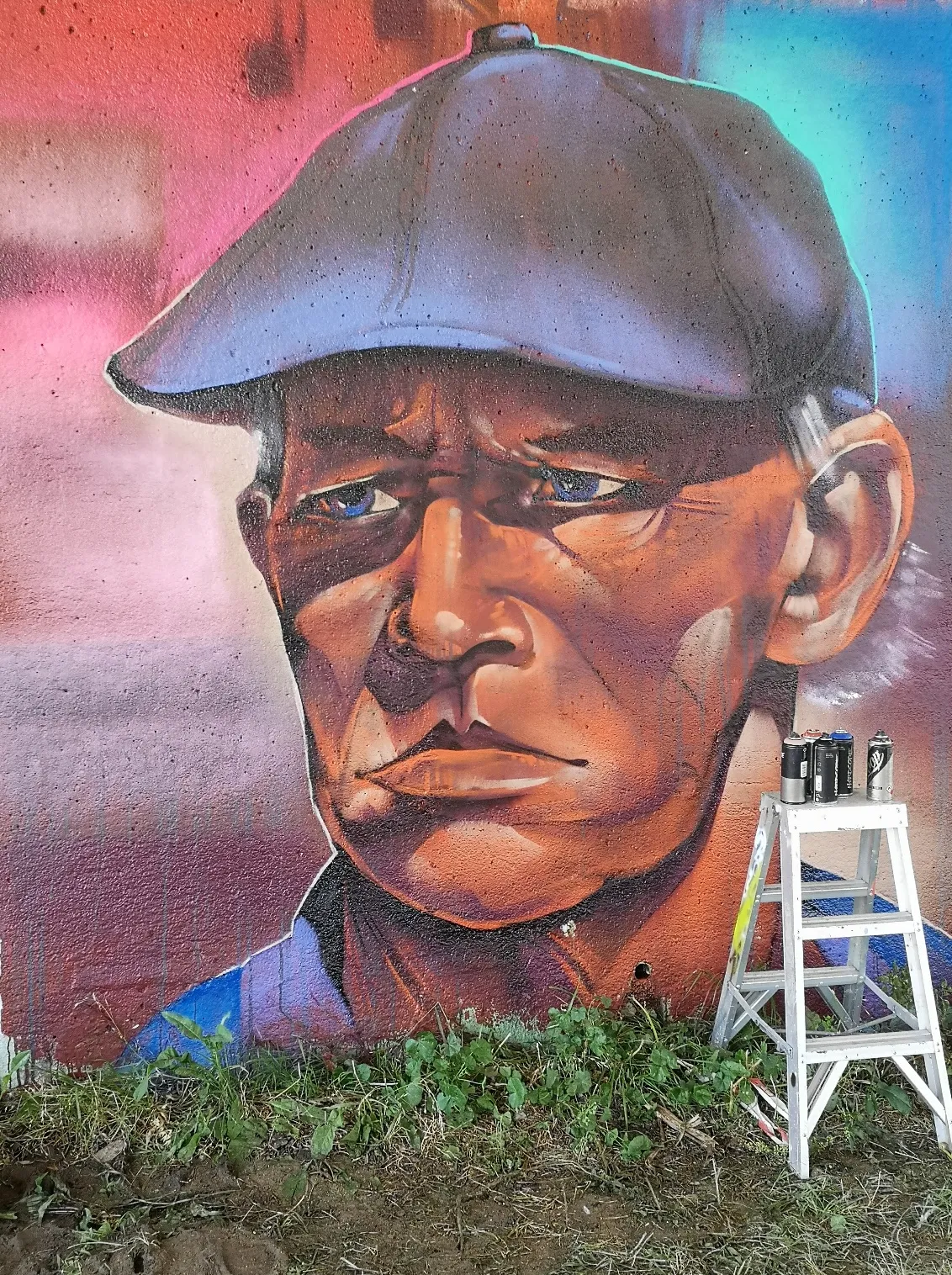 Realist portrait mural of a weathered man in a flat cap, painted with a ladder and spray cans still in front of it