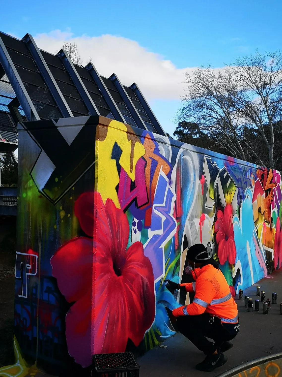 Artist in hi-vis kneeling to paint a large flower on a colourful graffiti wall